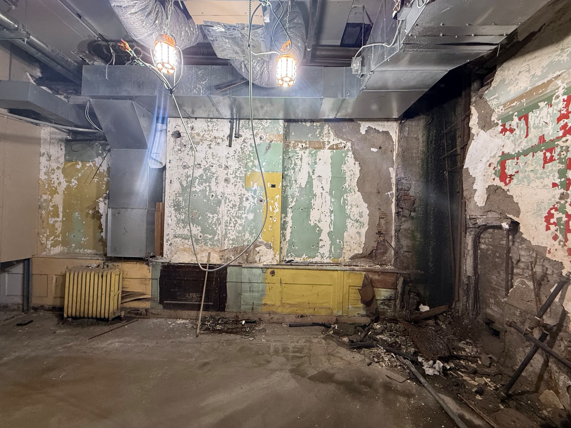 Basement room before construction with peeling multi-colored paint, exposed brick, overhead ductwork and temporary work lights.