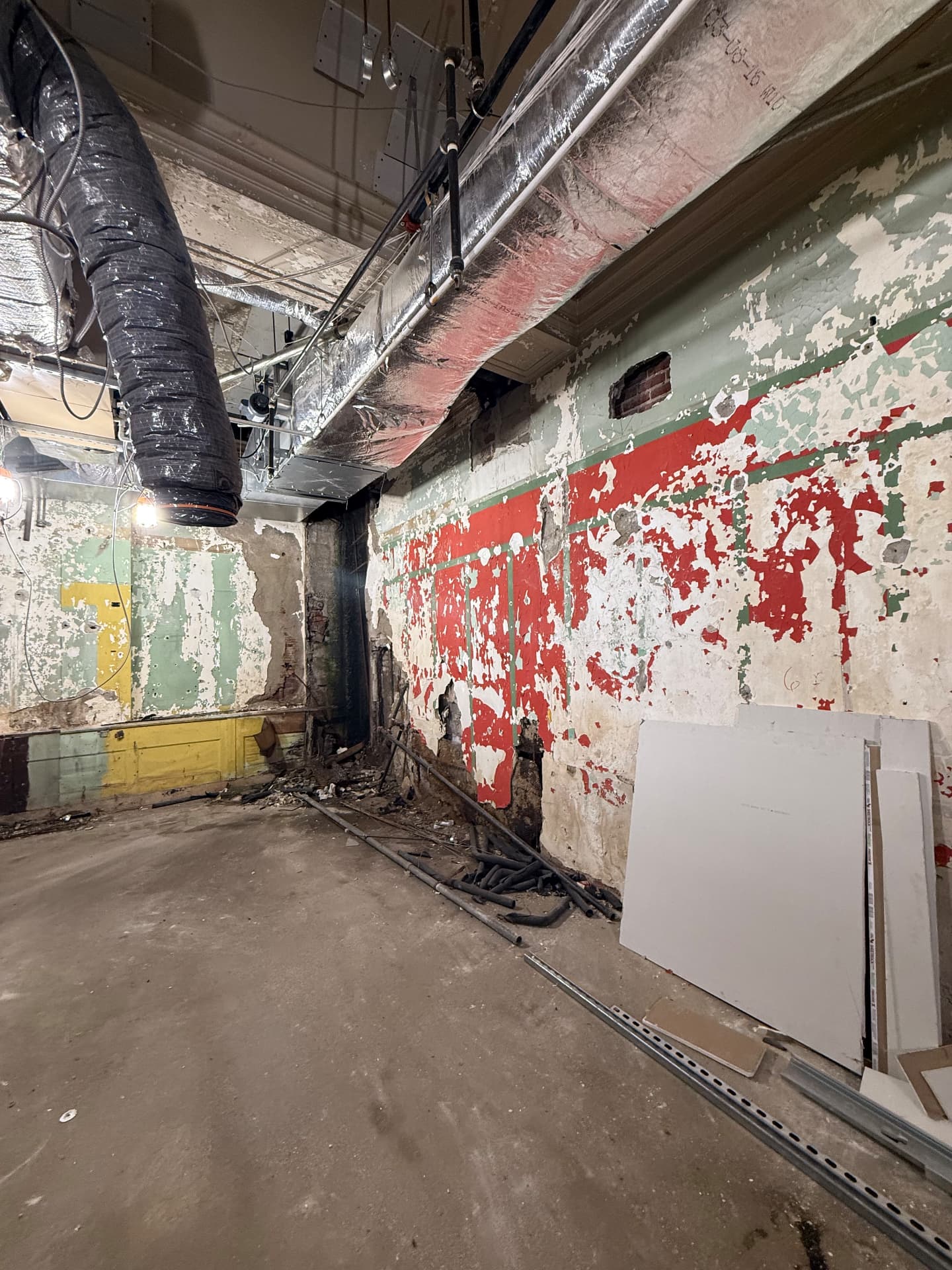 Basement corner before construction showing damaged painted walls, insulated ductwork, loose pipe and drywall sheets staged against the wall.