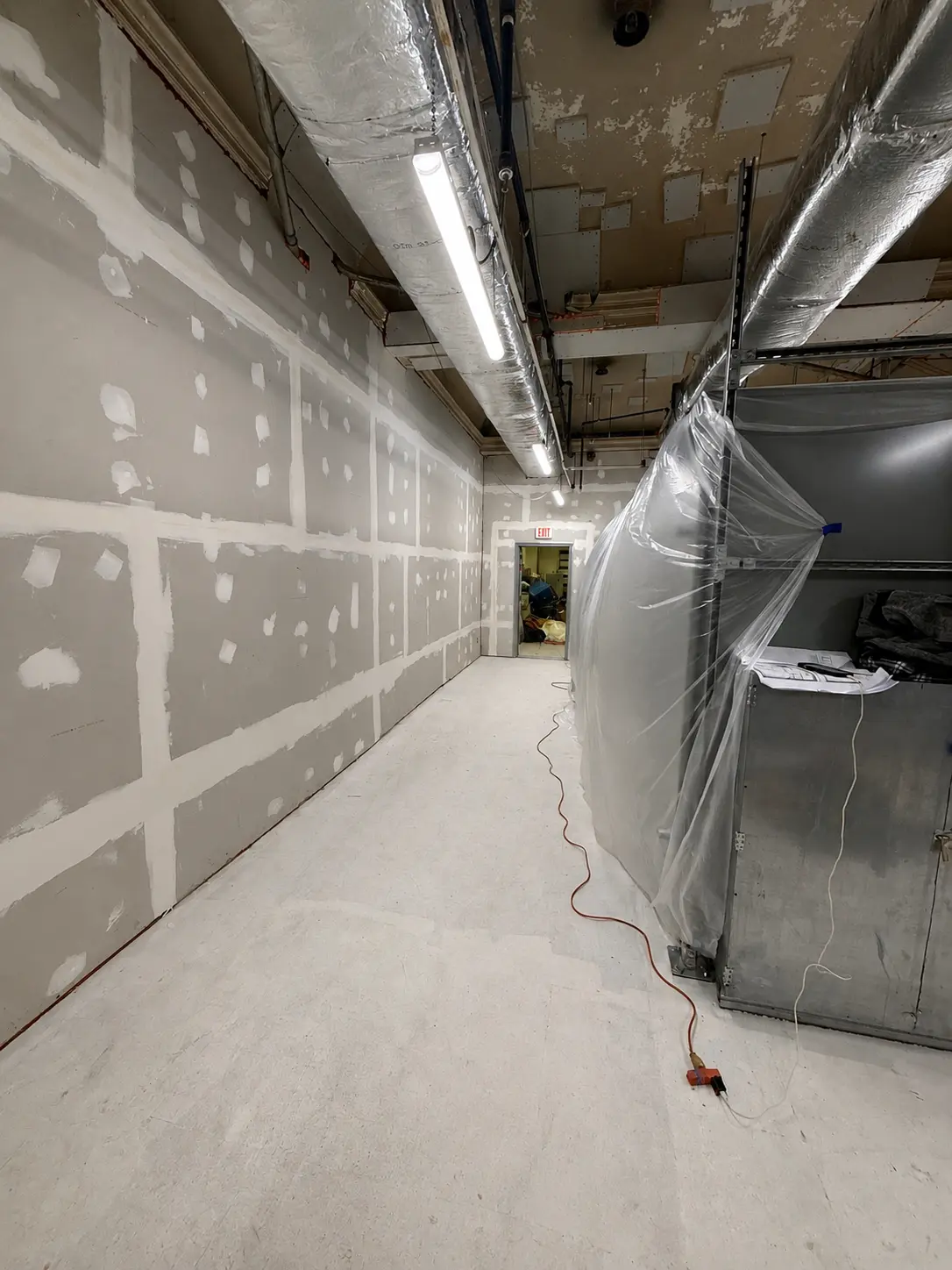 Basement equipment room during early construction — taped drywall, overhead ductwork and conduit, existing equipment wrapped in protective plastic.