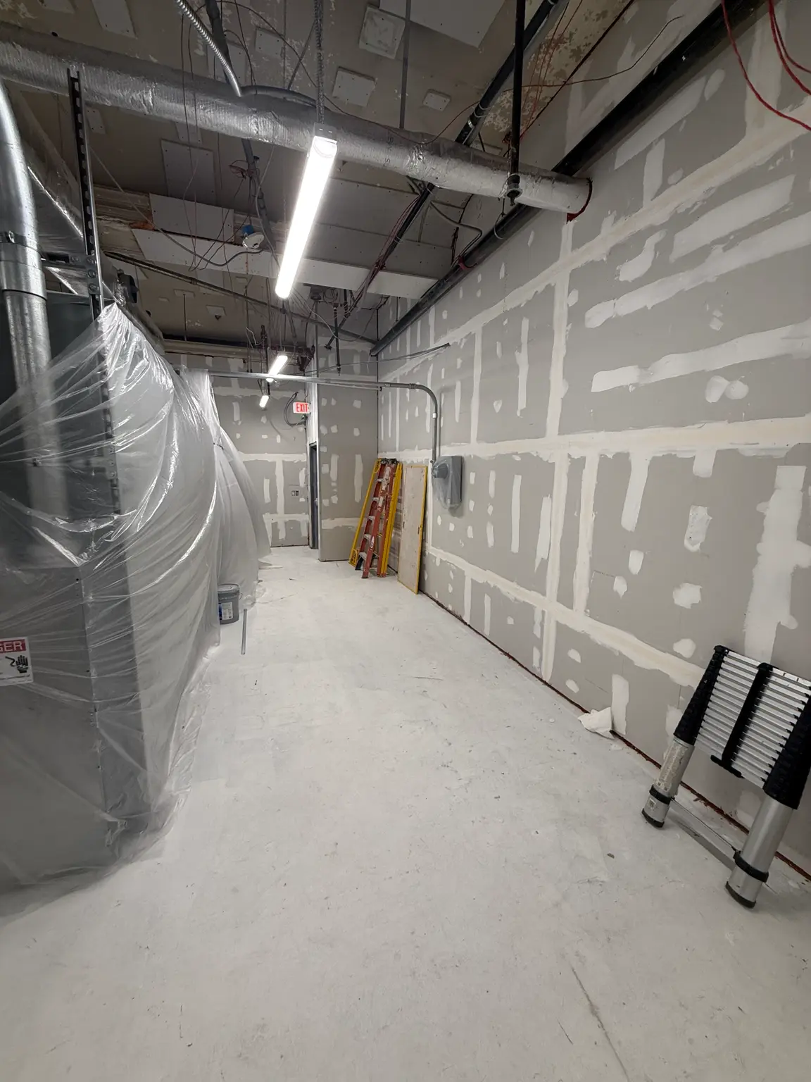 Corridor with fire-rated drywall assemblies taped and compounded, overhead conduit and piping, ladder and materials staged on site.