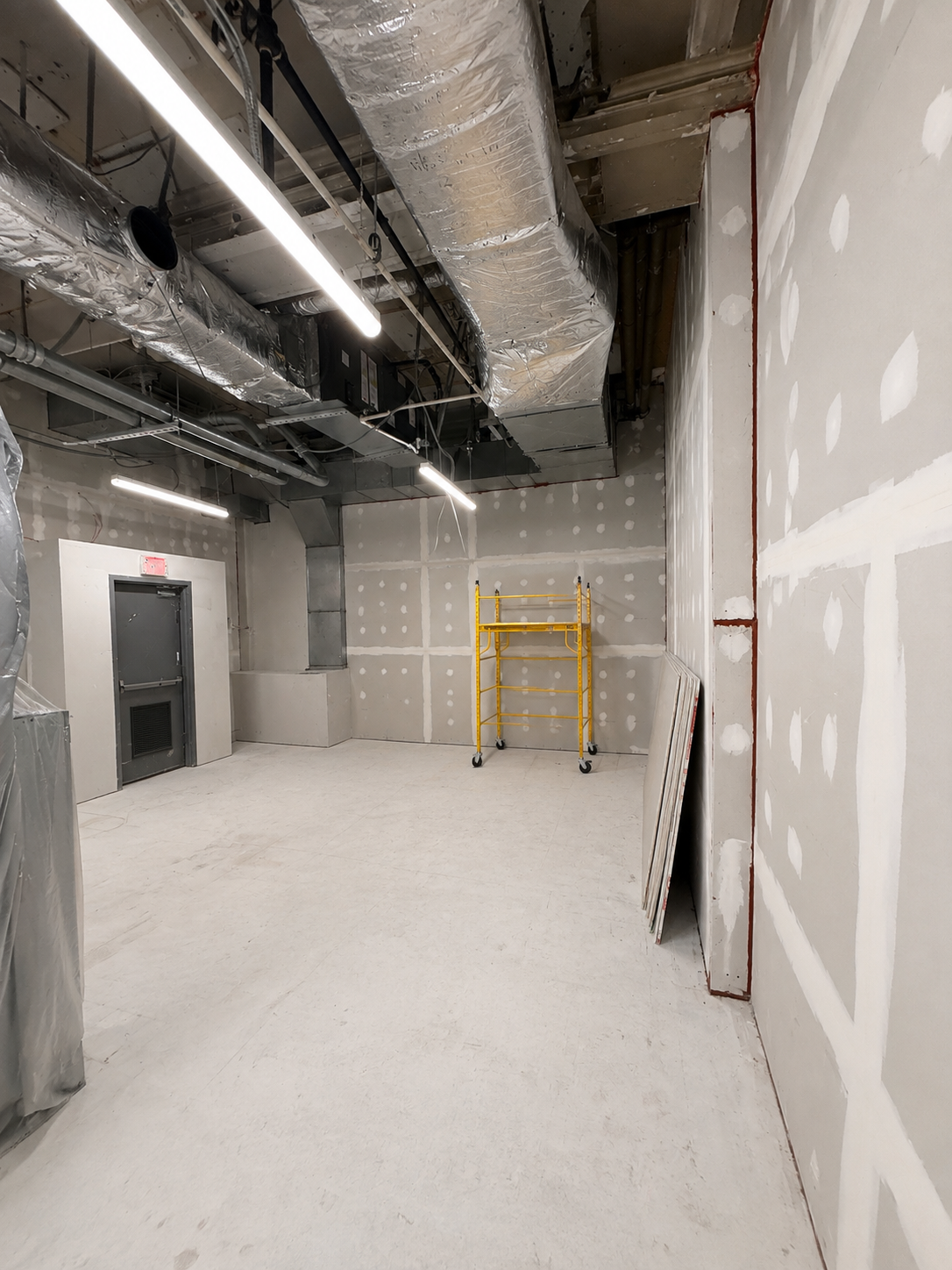 Basement room mid-construction with taped drywall walls, scaffold, insulated ductwork and a rated door opening.